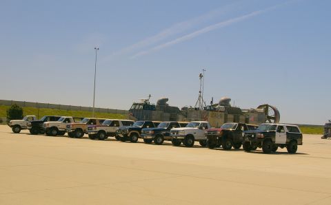1986 Ford Bronco - Several Ford trucks and Broncos are lined up on a paved surface with hovercrafts in the background under a clear blue sky.