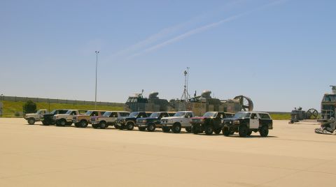 1986 Ford Bronco - Several SUVs and trucks are parked in a line on an airfield, with hovercraft visible in the distance.