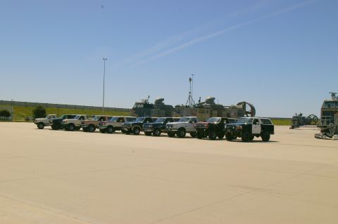 1986 Ford Bronco - A row of SUVs and trucks are parked outdoors under a clear blue sky with large boats in the distance.