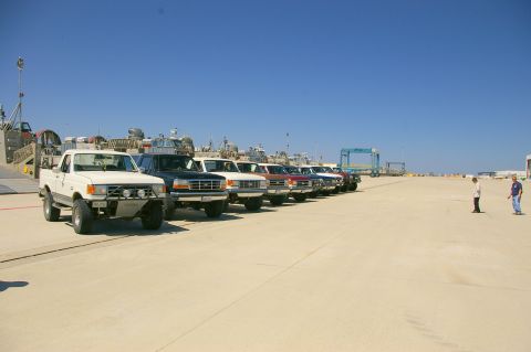 1986 Ford Bronco - A row of Ford trucks and SUVs are parked on a sunny day near a body of water with boats in the background.