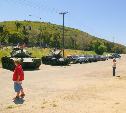 1986 Ford Bronco - A sunny day scene features two tanks and a row of pickup trucks parked along a road with a grassy hill in the background.