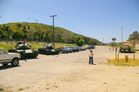 1986 Ford Bronco - A sunny outdoor scene shows two military tanks parked next to a row of civilian vehicles.