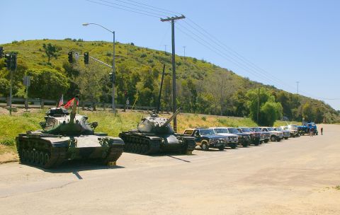 1986 Ford Bronco - Military tanks and a line of pickup trucks are parked roadside with a grassy hill in the background.