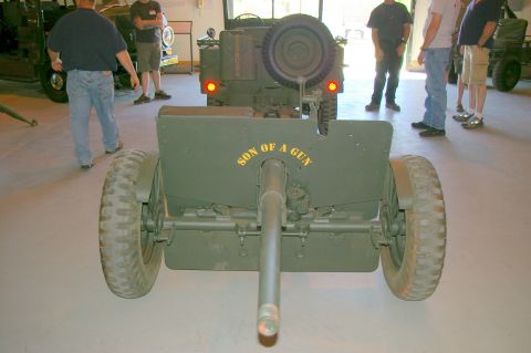 1986 Ford Bronco - Military artillery piece with painted text is displayed indoors near other vehicles and onlookers.