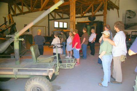 1986 Ford Bronco - A group of people observe a military vehicle inside a building, with other vehicles visible in the background.