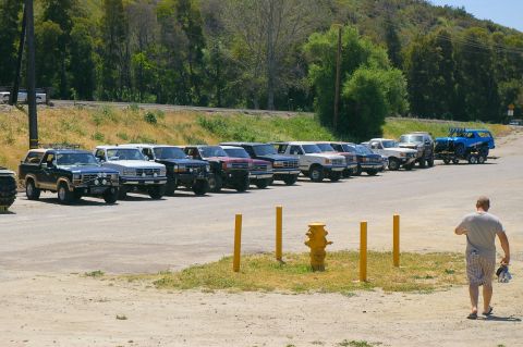 1986 Ford Bronco - A line of SUVs and trucks are parked in a lot with a man walking away from the camera.