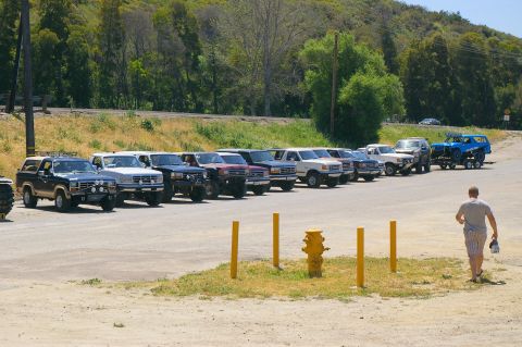1986 Ford Bronco - A row of Ford Broncos and other SUVs are parked in a lot, with a man walking away from the camera.