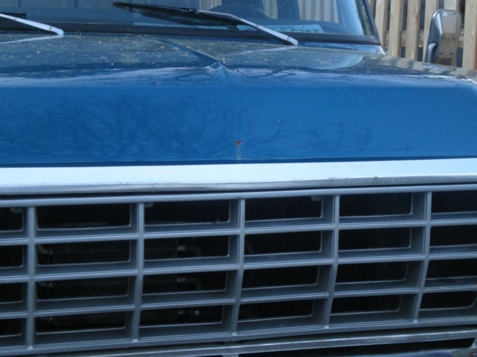 Close-up of a blue truck's grille and hood, showing horizontal bars on the grille and a dent on the hood