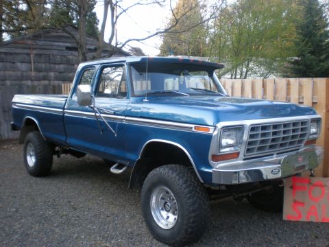 1993 Ford Bronco - A blue pickup truck is parked in a gravel lot with a wooden fence and building in the background.
