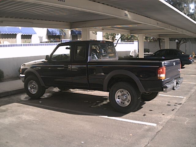 Dark Ford Ranger extended-cab pickup parked under a carport, seen from driver's side rear