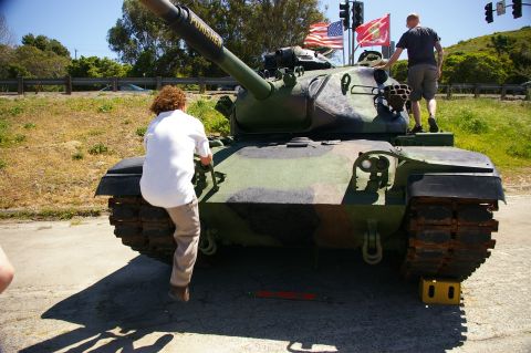 1986 Ford Bronco - A military tank with camouflage paint is shown with two people interacting with it.