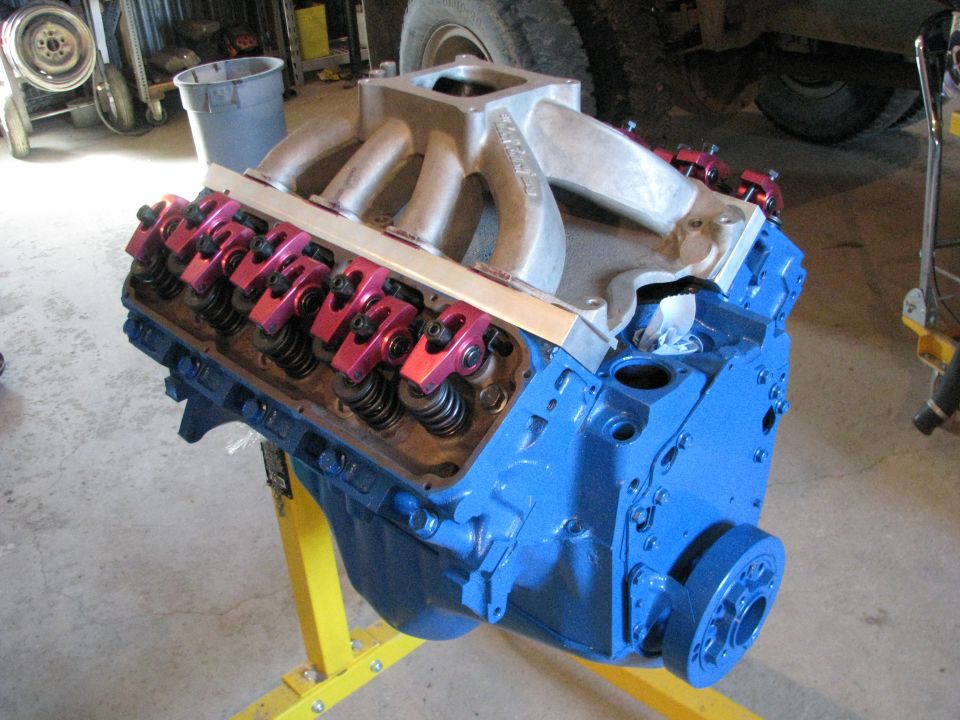 Blue V8 engine on a yellow stand, showing aluminum intake manifold and red roller rockers