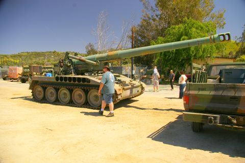 1986 Ford Bronco - A large military tank is parked on a dirt lot next to a camouflage pickup truck.