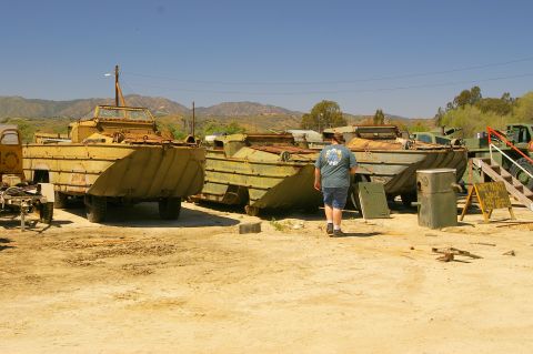 1986 Ford Bronco - Amphibious military vehicles are parked in a dusty lot under a clear blue sky.
