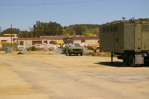 1986 Ford Bronco - A green pickup truck is parked in front of a building with a large trailer to its right.