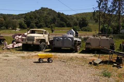 1986 Ford Bronco - Several military vehicles are parked in a lot with a small yellow wagon in the foreground.