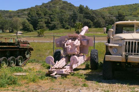 1986 Ford Bronco - An artillery piece and a military truck are parked in a yard with a fence and trees.