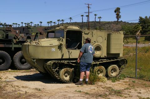 1986 Ford Bronco - A man stands beside a tracked cargo vehicle, with another military truck visible in the background under a clear sky.
