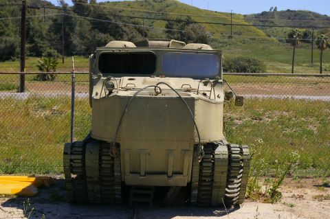1986 Ford Bronco - A military-style tracked vehicle sits parked outdoors behind a chain-link fence.