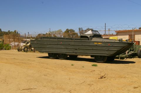 1986 Ford Bronco - An olive drab amphibious military vehicle is parked outdoors on a sandy lot, with a fence and buildings in the background.