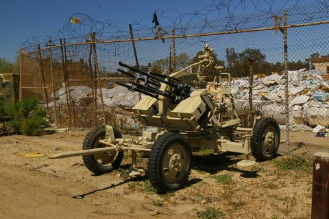 1986 Ford Bronco - A towed anti-aircraft gun is parked outdoors near a fence and a large pile of trash.