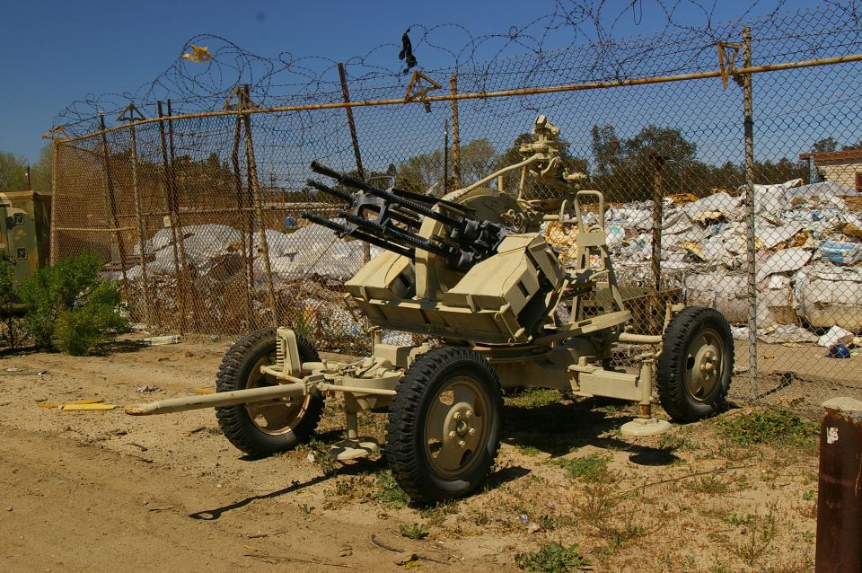 Wheeled anti-aircraft gun with multiple barrels, set against a chain-link fence and pile of debris