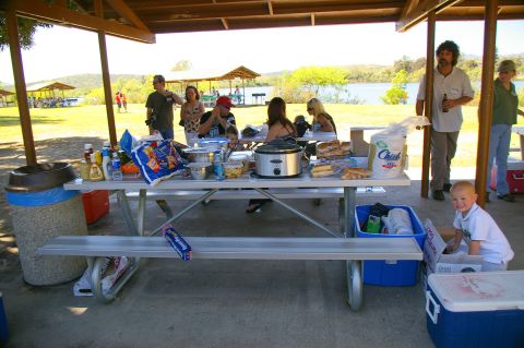 1986 Ford Bronco - A group enjoys a picnic under a shelter, with food laid out on a table and a lake visible in the distance.