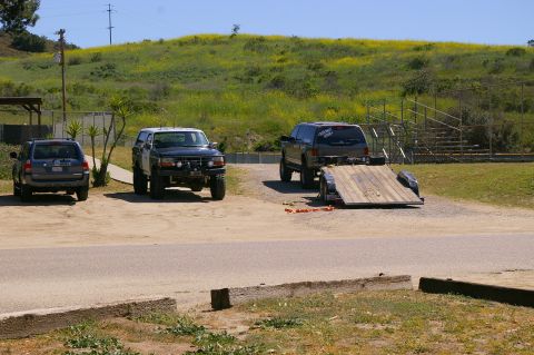 1986 Ford Bronco - Three SUVs and a trailer are parked in an outdoor lot with a grassy, yellow-flowered hill behind them.