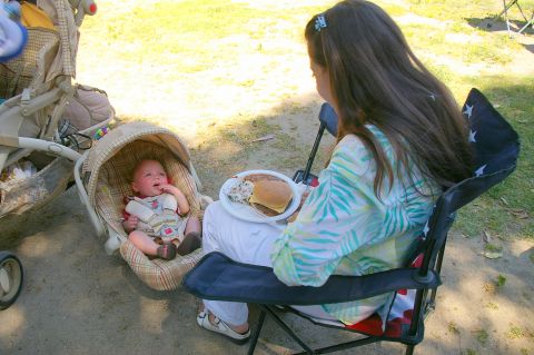 1986 Ford Bronco - A baby sits in a car seat next to a stroller while a woman eats a meal outdoors.