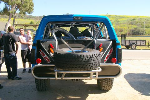 1986 Ford Bronco - The rear of a modified blue SUV is shown, a spare tire and roll cage, with people standing around it.
