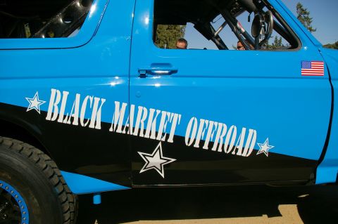 1986 Ford Bronco - This blue off-road truck features "BLACK MARKET OFFROAD" graphics and an American flag decal on the door.