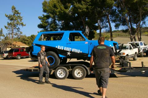 1986 Ford Bronco - A blue 1986 Ford Bronco, modified for off-road use, is loaded onto a trailer with other vehicles in the background.