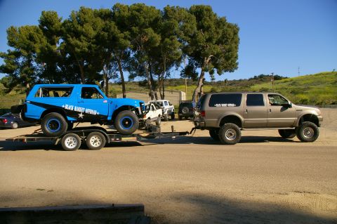 1986 Ford Bronco - A tan SUV tows a blue off-road SUV on a trailer, with other vehicles visible in the background.