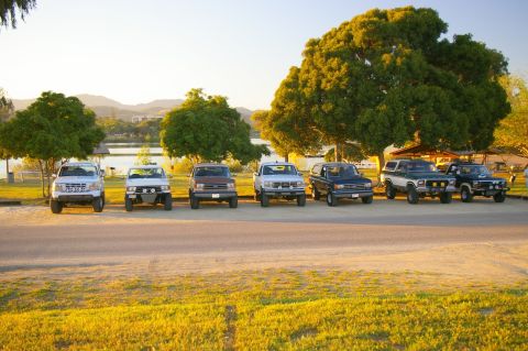 1986 Ford Bronco - Several SUVs and trucks are parked in a line near a lake during a golden hour sunset.