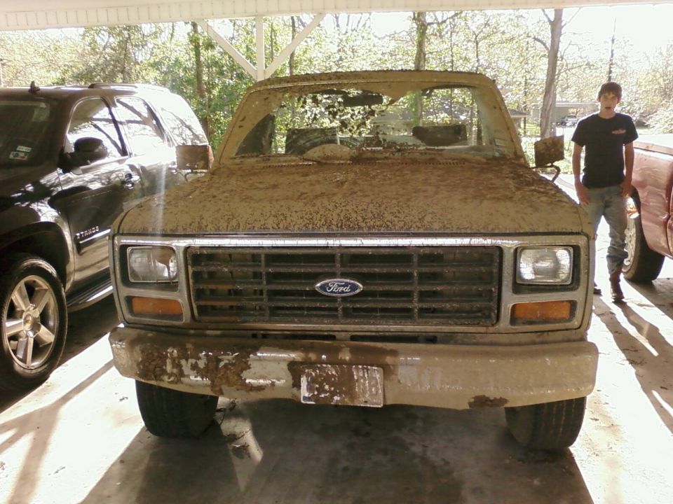 Mud-covered 1980s Ford truck front end, parked next to a black SUV and a red truck. A person stands to the right