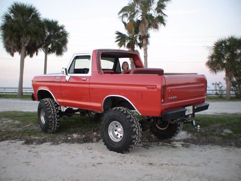 1978 Ford Bronco - A lifted red Ford Bronco with a white top is parked on a sandy area with palm trees in the background.