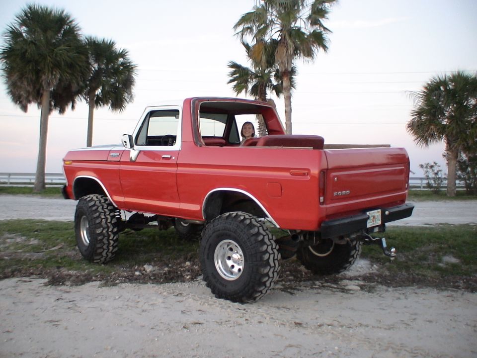 Red lifted 1978 Ford Bronco with white top parked on dirt near palm trees