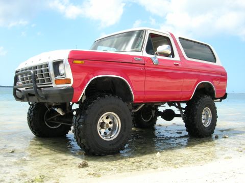1978 Ford Bronco - This lifted 1978 Ford Bronco, painted red and white, is parked on a beach with shallow water around its tires.