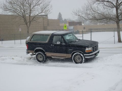 1996 Ford Bronco - A black and tan Ford Bronco is stuck in the snow on a street with a school in the background.