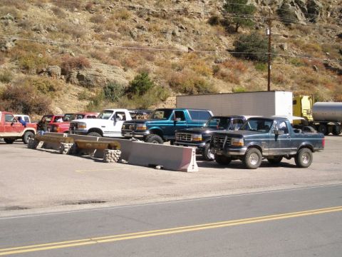 1996 Ford Bronco - A lineup of Ford Broncos and trucks are parked in a lot, with a hillside visible behind them.