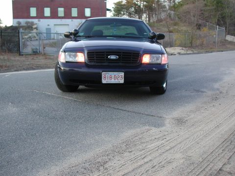 1999 Ford Crown Victoria - A dark blue Ford Crown Victoria is parked on a road in front of a brick building.