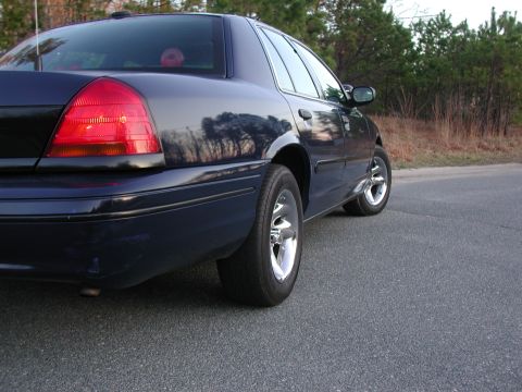 1999 Ford Crown Victoria - A dark blue sedan is viewed from the rear quarter angle on a paved road.