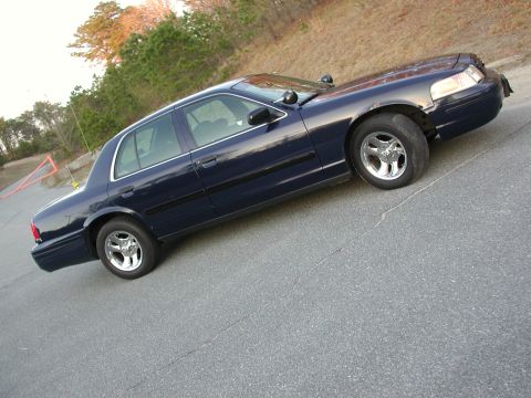 1999 Ford Crown Victoria - A dark blue sedan is parked on a paved surface with trees in the background.
