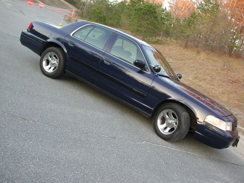 1999 Ford Crown Victoria - A dark blue sedan, likely a 1998 model, is parked on a cracked asphalt road with trees in the background.