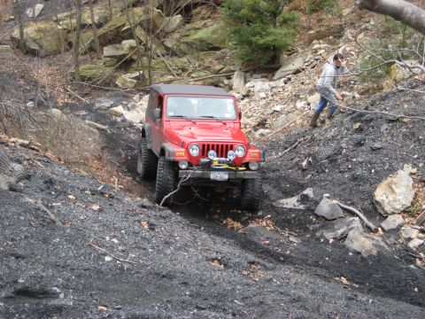 1991 Ford Bronco - Red Jeep Wrangler navigates a challenging off-road trail on a steep, rocky incline.