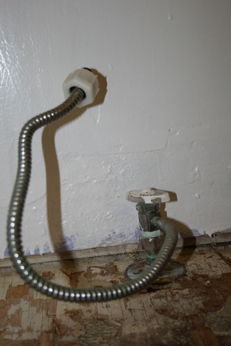 Two flexible metal water supply lines connected to shut-off valves on a wall above damaged wooden flooring