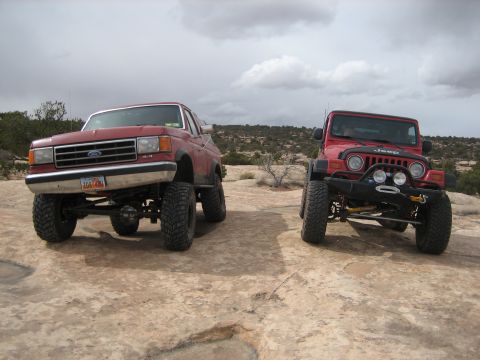 1990 Ford Bronco - Two modified off-road vehicles, a Ford pickup and a Jeep, are parked on a rocky desert landscape.