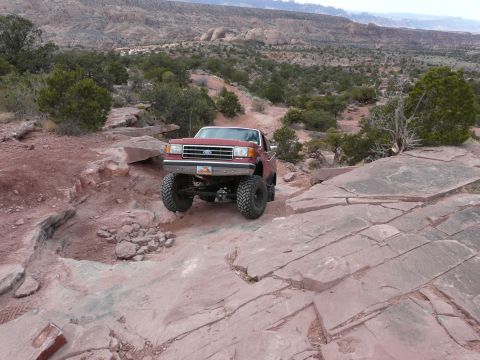 1990 Ford Bronco - A lifted Ford pickup truck tackles a challenging off-road path with a vast desert landscape in the background.