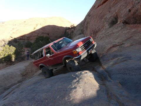 1990 Ford Bronco - A red Ford Bronco navigates a steep, rocky incline under a clear sky.