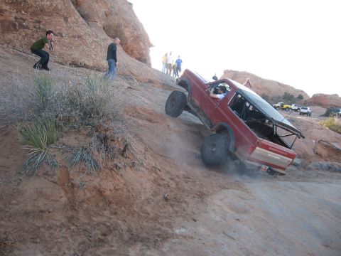 1990 Ford Bronco - A modified red SUV navigates a challenging off-road trail with spectators observing the ascent.